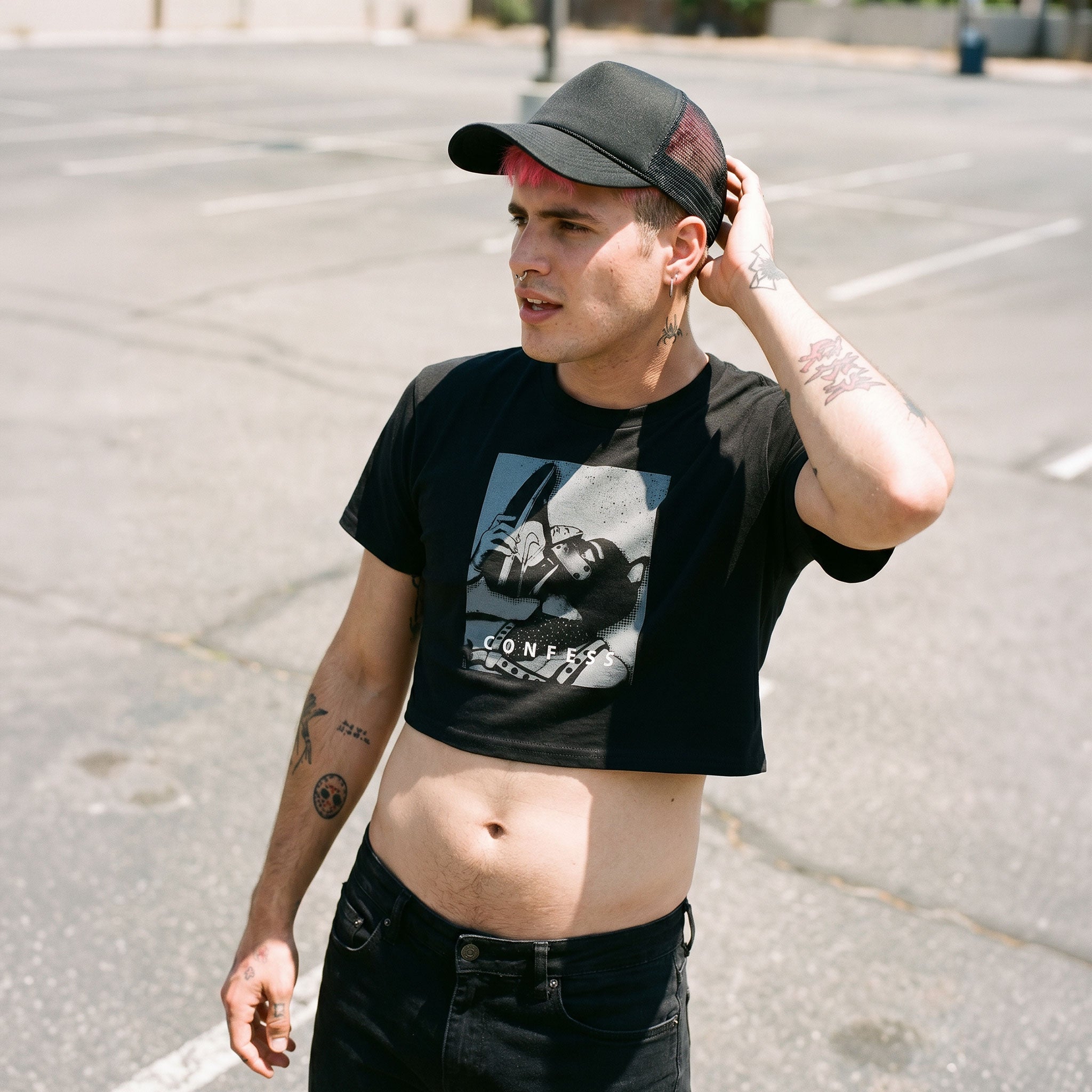 Gay Man wearing a Confess Crop Sniff Test, a black pup graphic crop top and a cap, standing on a street.