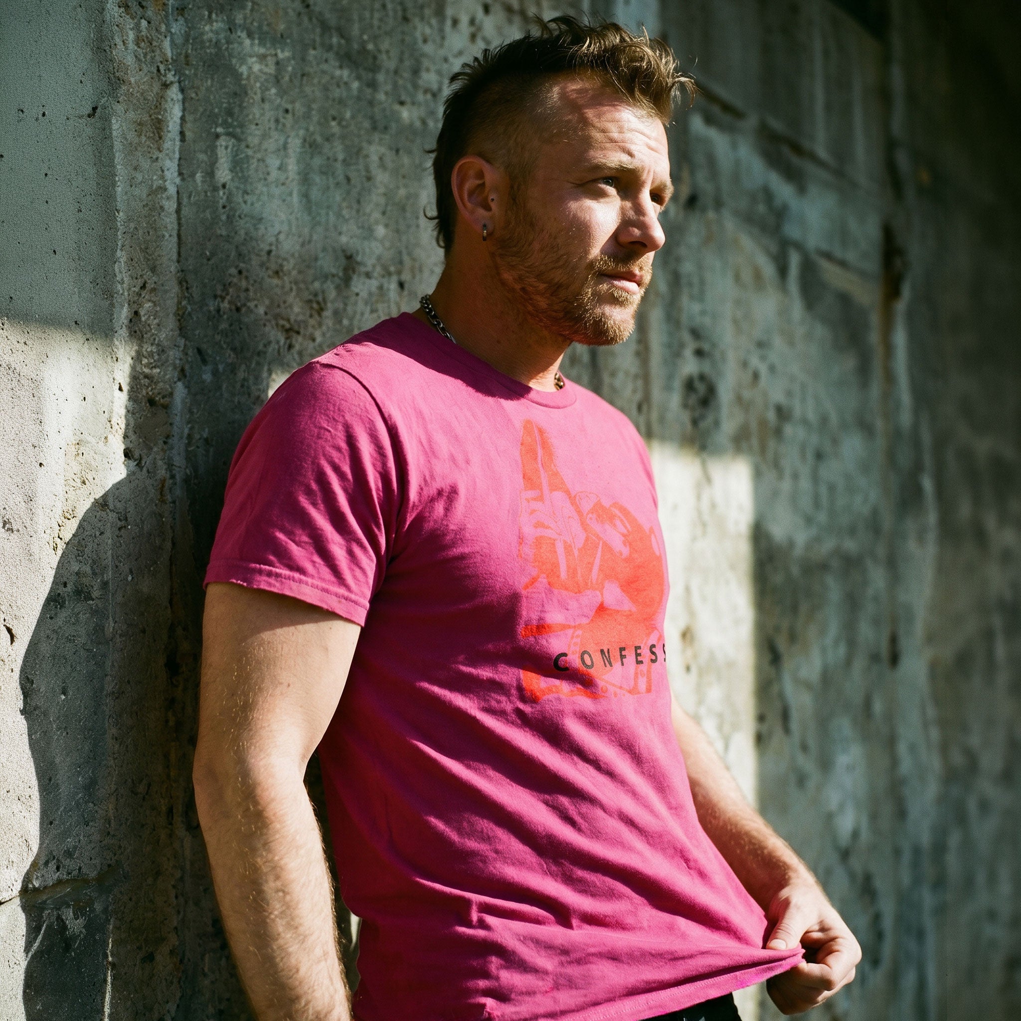 Lifestyle shot of a gay man wearing a berry pink t-shirt with a Sneaker Pup graphic design and 'CONFESS' text standing in front of a concrete wall