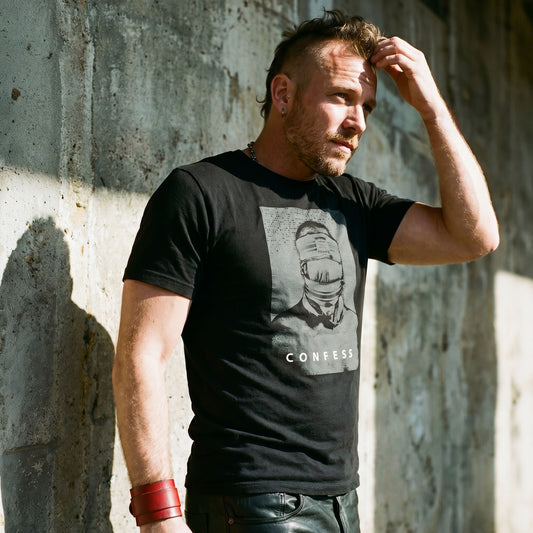 Lifestyle image of A gay man wearing a black t-shirt with a Blind Jock graphic design and 'CONFESS' text in front of a concrete wall.