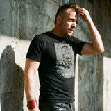 Lifestyle image of A gay man wearing a black t-shirt with a Blind Jock graphic design and 'CONFESS' text in front of a concrete wall.