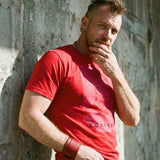 Lifestyle image of A gay man wearing a red t-shirt with a Blind Jock graphic design and 'CONFESS' text in front of a concrete wall.