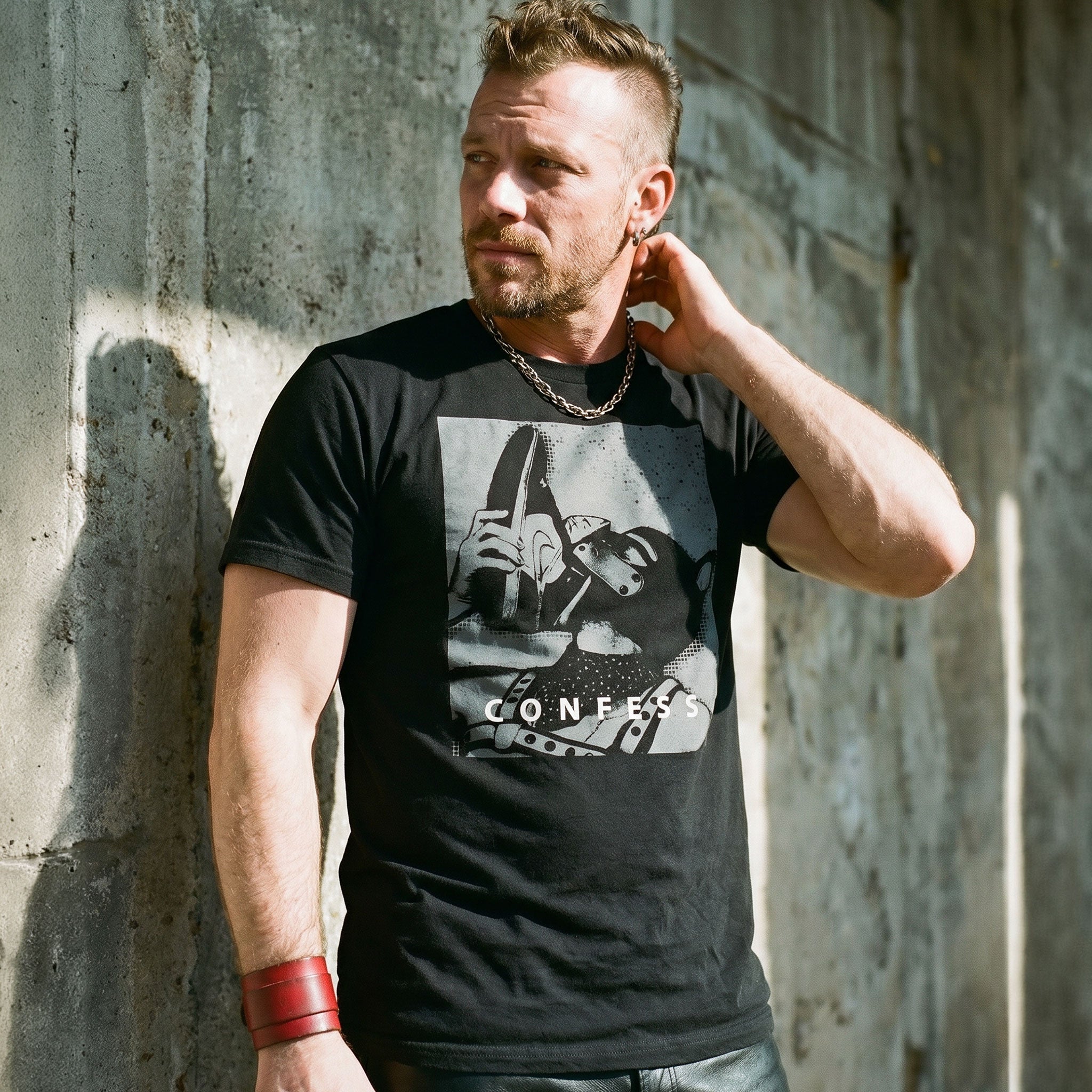 Lifestyle shot of a gay man wearing a black t-shirt with a Sneaker Pup graphic design and 'CONFESS' text standing in front of a concrete wall