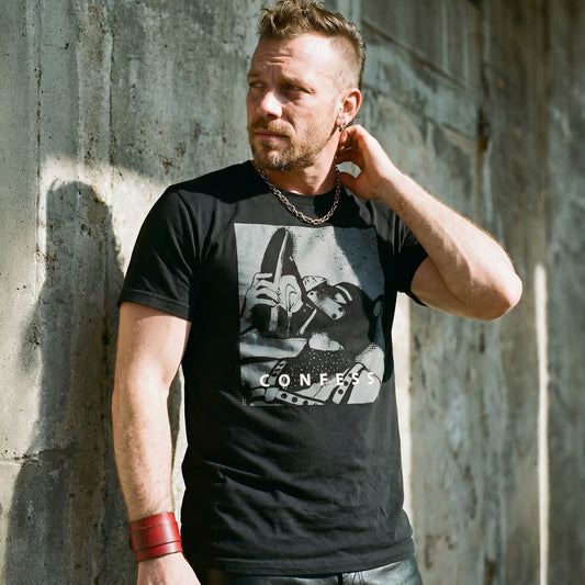 Lifestyle shot of a gay man wearing a black t-shirt with a Sneaker Pup graphic design and 'CONFESS' text standing in front of a concrete wall