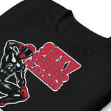 Folded black t-shirt with a man in rubber with red helmet and shoes sitting on top of a square barrel of lube graphic design on a beige background  - Black and red Sexy Rubber Flanders t-shirt - kinky latex rubber kink tee by BackRoomGear